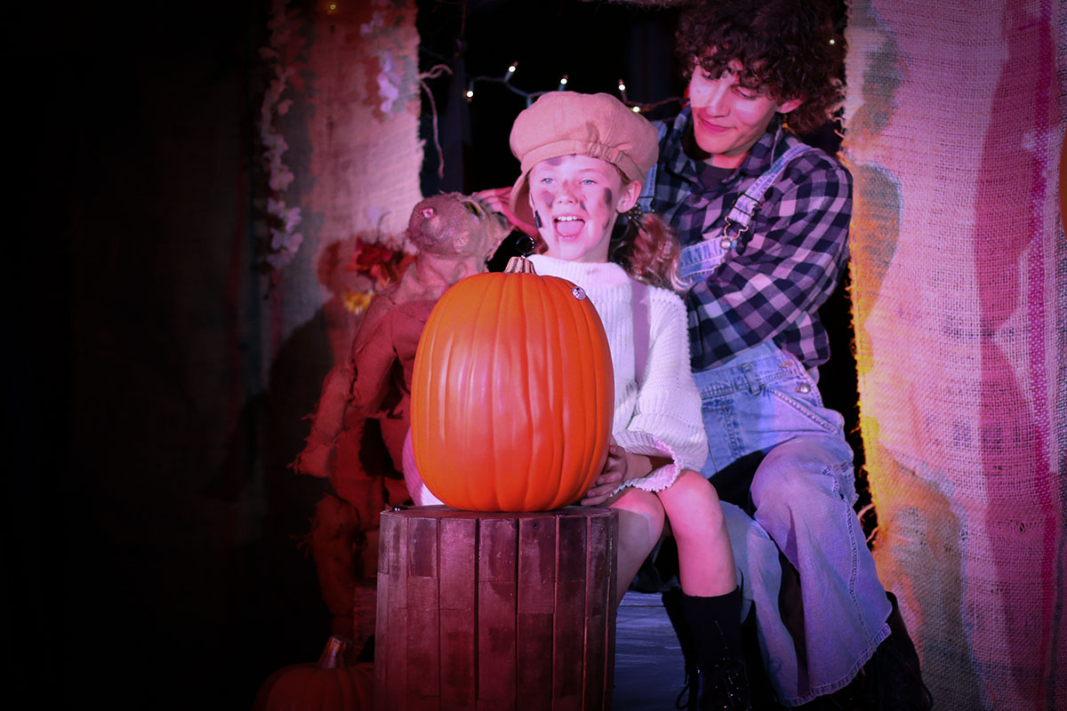 Jane Curry as the Pumpkin Kid seated behind a glowing pumpkin on a wooden barrel